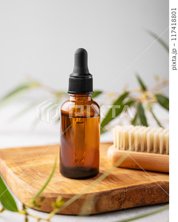 Nail oil or serum in a bottle on a wooden board with a brush on a gray background with green leaves. Nail oil or serum in a bottle on a wooden board with a brush on a gray background with green leaves. 117418801