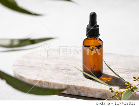 Face and body serum in bottle on a marble board on a white background with green leaves and shadow. 117418813