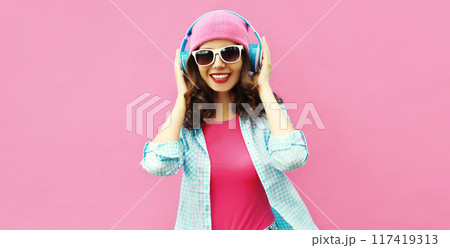 Colorful portrait of happy young woman listening to music in headphones in hat on pink background 117419313