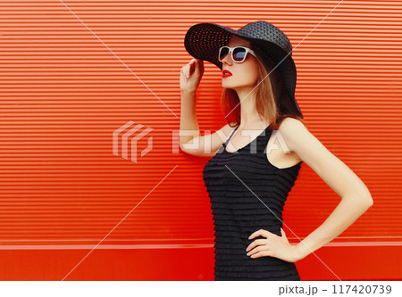 Portrait of beautiful young woman wearing a black summer hat and dress on red background Portrait of beautiful young woman wearing a black summer hat and dress on red background 117420739