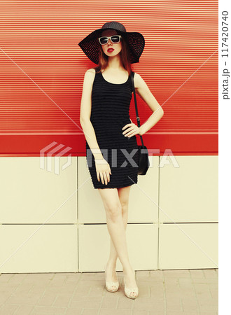 Portrait beautiful young woman with handbag wearing a black summer hat and dress on red background Portrait beautiful young woman with handbag wearing a black summer hat and dress on red background 117420740