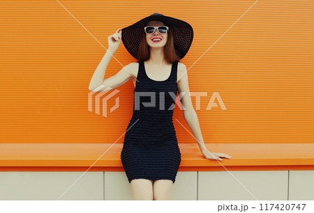 Portrait of beautiful young woman wearing a black summer hat and dress on orange background Portrait of beautiful young woman wearing a black summer hat and dress on orange background 117420747