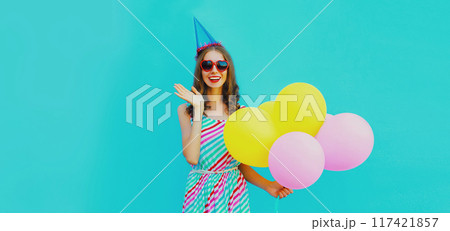 Beautiful happy cheerful young woman with balloon in birthday cap in bright dress on blue background Beautiful happy cheerful young woman with balloon in birthday cap in bright dress on blue background 117421857