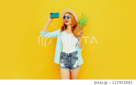 Happy smiling woman with pineapple taking selfie with phone in summer hat on color yellow background Happy smiling woman with pineapple taking selfie with phone in summer hat on color yellow background 117422681