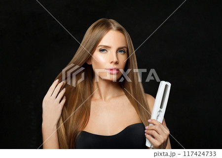 Beautiful brunette woman holding straight hair iron and straightening her long smooth shiny hair  117424733