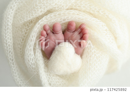 The tiny foot of a newborn baby. Soft feet of a new born in a white wool blanket. Close up of toes, heels and feet of a newborn. Knitted white heart in the legs of a baby. Macro photography.  117425892
