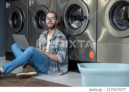 Man multitasking with laptop and household chores sitting on floor waiting at self-service laundry Man multitasking with laptop and household chores sitting on floor waiting at self-service laundry 117427077