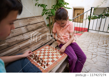 Children playing chess outside on a rustic wooden bench in a courtyard, focused on the game and strategizing moves in a calm and relaxed setting. Children playing chess outside on a rustic wooden bench in a courtyard, focused on the game and strategizing moves in a calm and relaxed setting. 117427274