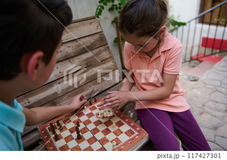 Children engaging in a thoughtful outdoor chess game on a wooden bench, showcasing strategy, concentration, and friendly competition in an informal setting. Children engaging in a thoughtful outdoor chess game on a wooden bench, showcasing strategy, concentration, and friendly competition in an informal setting. 117427301