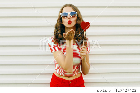 Portrait of beautiful young woman with red heart shaped sunglasses blowing kiss on white background 117427422