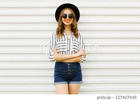 Happy smiling young woman in black round hat, shorts, white striped shirt on white wall background Happy smiling young woman in black round hat, shorts, white striped shirt on white wall background 117427430
