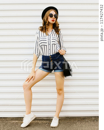 Pretty young woman posing in black round hat, shorts, white striped shirt posing on white background Pretty young woman posing in black round hat, shorts, white striped shirt posing on white background 117427432