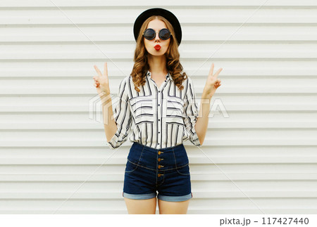 Attractive young woman blowing kiss in black round hat, shorts, striped shirt on white background 117427440