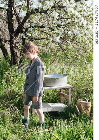 a little boy in shorts and a jacket waters plants from a watering can. Fashionable look for a country boy a little boy in shorts and a jacket waters plants from a watering can. Fashionable look for a country boy 117427957
