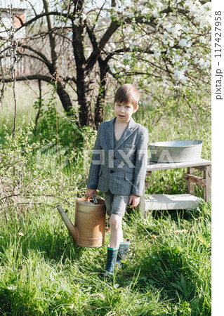 a little boy in shorts and a jacket waters plants from a watering can. Fashionable look for a country boy a little boy in shorts and a jacket waters plants from a watering can. Fashionable look for a country boy 117427958