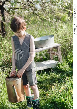 little boy in shorts and a vest waters plants from a watering can. Fashionable look for a country boy little boy in shorts and a vest waters plants from a watering can. Fashionable look for a country boy 117427959