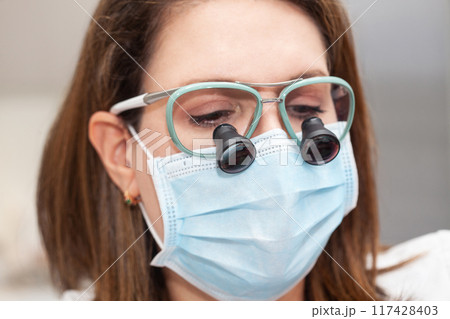 Close up of a female dermatologist face while suturing a patient during a small surgery wearing a binocular loupe. Close up of a female dermatologist face while suturing a patient during a small surgery wearing a binocular loupe. 117428403