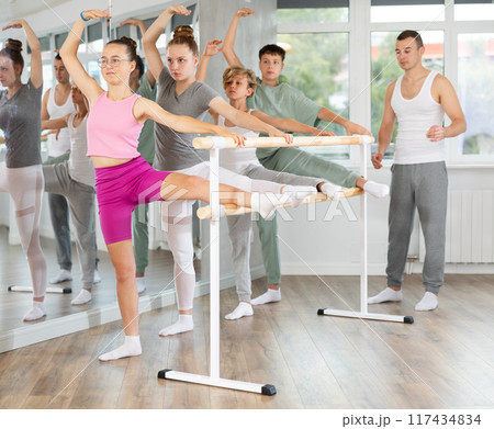 Teen ballet students prepare for classes, near ballet bare stretch legs under man teacher guidance Teen ballet students prepare for classes, near ballet bare stretch legs under man teacher guidance 117434834