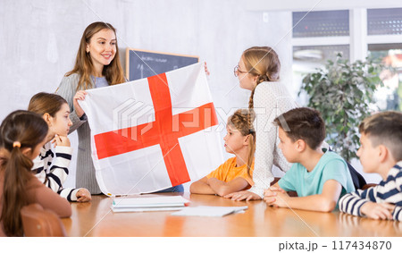 Young female teacher showing flag of England to schoolchildren preteens during history lesson in classroom 117434870