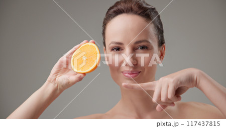 Beauty, health, cosmetics, anti-aging therapy and skin care concept - young beautiful brunette Caucasian woman holding half a orange, concept of freshness, vitamins and nutrients 117437815