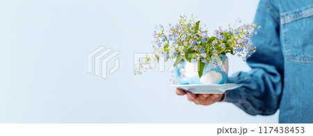 Hand holding blue cup with white cloud patterns, filled with flowers. 117438453