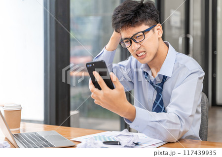 Frustrated and angry young businessman working late at the office trying to finish his project holding a phone in one hand looking angry at the screen Frustrated and angry young businessman working late at the office trying to finish his project holding a phone in one hand looking angry at the screen 117439935