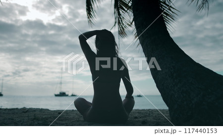 Young woman enjoying a peaceful moment on a tropical beach at sunset, surrounded by palm trees and boats 117440153