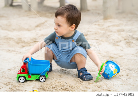 Little cute toddler boy 2.5 years old plays in the sandbox on a sunny summer day. Outdoor creative activities for kids. Toy car and shovel. 117442902