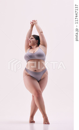 Chubby young lady posing shirtless showing her confidence against white studio background. Model dressed grey underwear. Chubby young lady posing shirtless showing her confidence against white studio background. Model dressed grey underwear. 117447137
