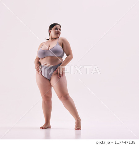 Full length photo of young overweight lady, model plus size posing in grey underwear highlights her shapes against white studio background. 117447139