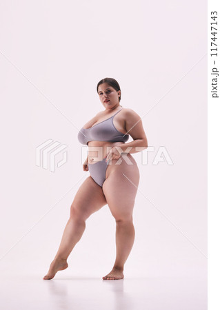 Chubby young lady posing shirtless showing her confidence against white studio background. Model dressed grey lingerie. 117447143