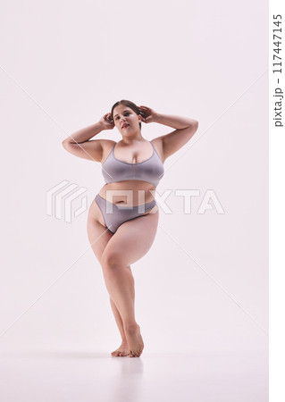Full length photo of young overweight lady, model plus size posing in grey lingerie highlights her shapes against white studio background. 117447145