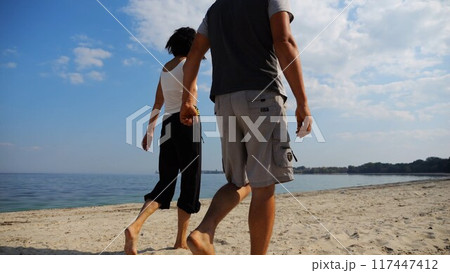 Couple holding hands of each other and walking at coast. Happy pair going along seashore at sunny day. Lovers spending time together at resort. Concept of vacation or holiday. Slow mo Couple holding hands of each other and walking at coast. Happy pair going along seashore at sunny day. Lovers spending time together at resort. Concept of vacation or holiday. Slow mo 117447412