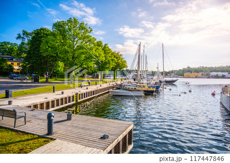 Sailboats moored at Skeppsholmen Harbor against a backdrop of clear skies with sunlight bathing the tranquil waters and tree-lined promenade. Sailboats moored at Skeppsholmen Harbor against a backdrop of clear skies with sunlight bathing the tranquil waters and tree-lined promenade. 117447846