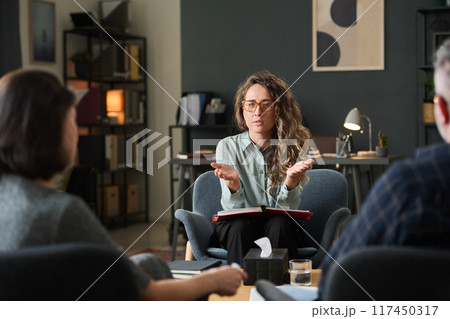 Three colleagues engaging in discussion about work strategies in a modern office setting, collaborating for project advancement and sharing ideas, seated around the table, Caucasian woman talking 117450317