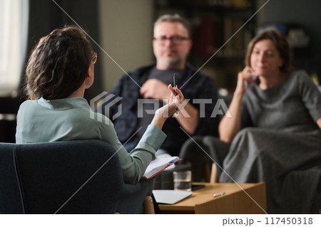 Couple having therapy session with female therapist gesturing while holding pen, room with bookshelf and therapy materials, focus on conversation, atmosphere calm and professional Couple having therapy session with female therapist gesturing while holding pen, room with bookshelf and therapy materials, focus on conversation, atmosphere calm and professional 117450318