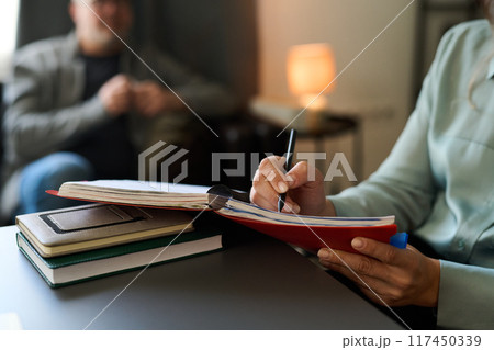 Hand writing notes in red notebook during counseling session with client in blurred background taking place in cozy, dimly lit room with books on table 117450339