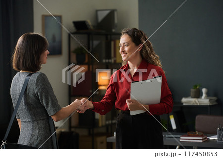 Two businesswomen greeting each other with handshake in modern office setting symbolizing professional agreement. One holding clipboard while they stand near desk and shelves Two businesswomen greeting each other with handshake in modern office setting symbolizing professional agreement. One holding clipboard while they stand near desk and shelves 117450853