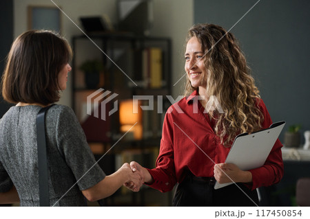 Professional businesswoman in red shirt greeting business colleague with handshake in office environment holding clipboard in curly hair Professional businesswoman in red shirt greeting business colleague with handshake in office environment holding clipboard in curly hair 117450854