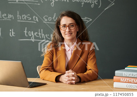 Front view portrait of adult woman as teacher looking at camera while sitting at desk in school with blackboard in background, copy space Front view portrait of adult woman as teacher looking at camera while sitting at desk in school with blackboard in background, copy space 117450855