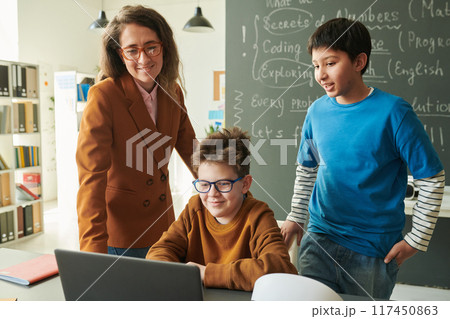 Portrait of smiling female teacher and two young boys looking at laptop screen in school classroom with blackboard in background 117450863