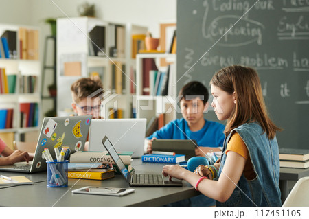 Side view portrait of young girl with prosthetic arm using laptop with inclusive group of children in school library copy space 117451105
