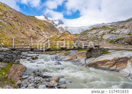 The clear waters of Schlaten Glacier stream rush over rocky beds beneath a wooden bridge, surrounded by the stunning Alpine landscape of Hohe Tauern National Park. 117451833
