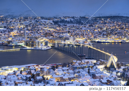 Panorama of Norwegian city of Troms in the winter. Snowy roofs, bridge, embankment near the port and fishing ships. Evening lights Panorama of Norwegian city of Troms in the winter. Snowy roofs, bridge, embankment near the port and fishing ships. Evening lights 117456557