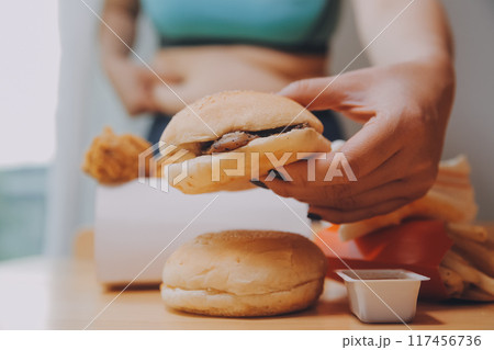 To eat or not. Cheerless plump woman looking at the burger while thinking to eat it or not 117456736