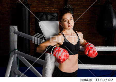 Fit strong brunette woman in sportswear top and shorts in boxing gloves in the ring looks at the camera Fit strong brunette woman in sportswear top and shorts in boxing gloves in the ring looks at the camera 117457482