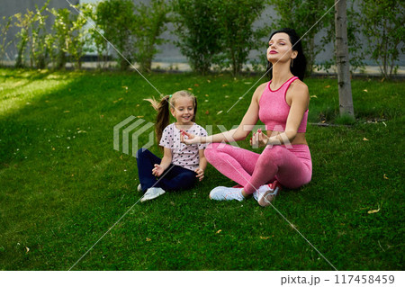 Woman and girl practicing yoga together outdoors on a sunny day in a green setting Woman and girl practicing yoga together outdoors on a sunny day in a green setting 117458459