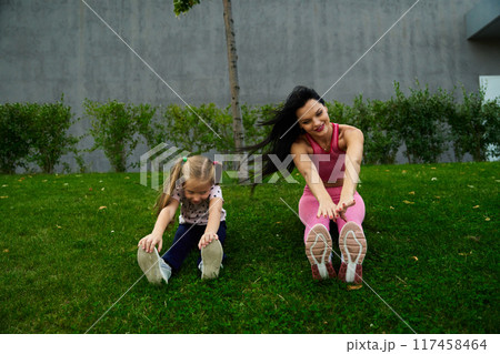 A woman and a girl doing stretching exercises on green grass in a park on a sunny day 117458464