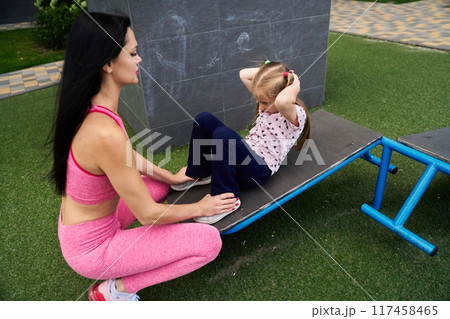 A mother guides her daughter in exercise at an outdoor fitness area during the afternoon 117458465