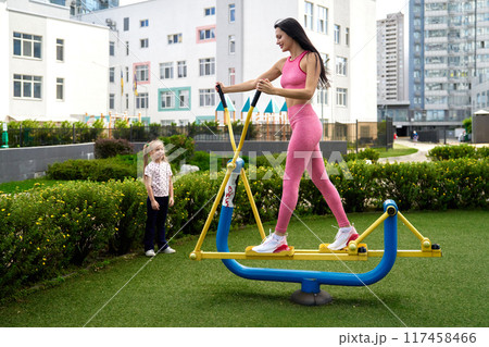 Woman exercising on outdoor fitness equipment while a child watches in a city park during daytime Woman exercising on outdoor fitness equipment while a child watches in a city park during daytime 117458466